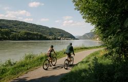 Zwei Radfahrende auf einem asphaltierten Uferweg entlang der Donau; gegenüber liegen bewaldete Hügel, rechts spenden Bäume Schatten.