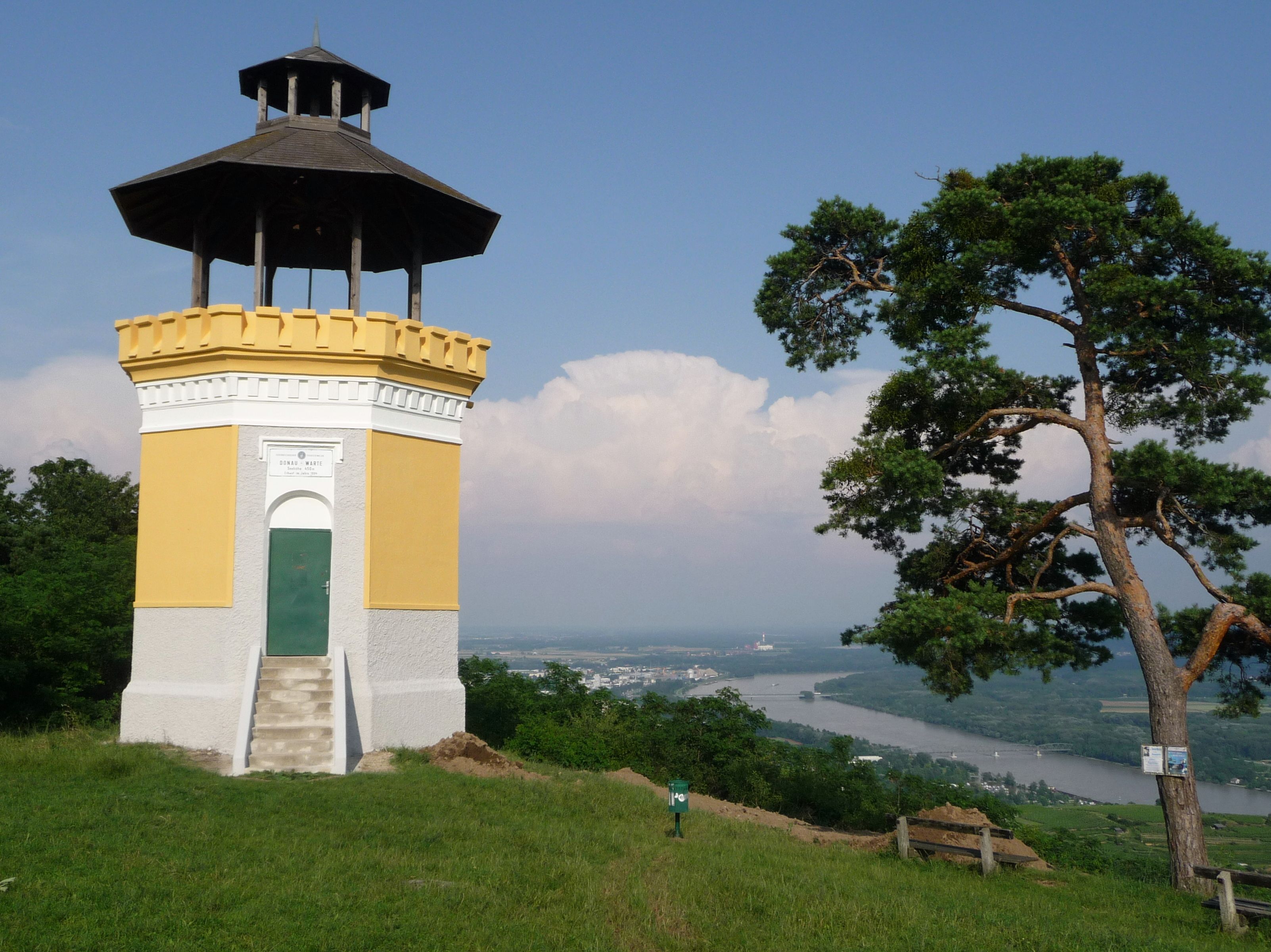 Ein gelber Aussichtsturm mit dunklem Dach steht auf einem Hügel mit Blick auf den Fluss Donau und eine Stadt im Hintergrund.