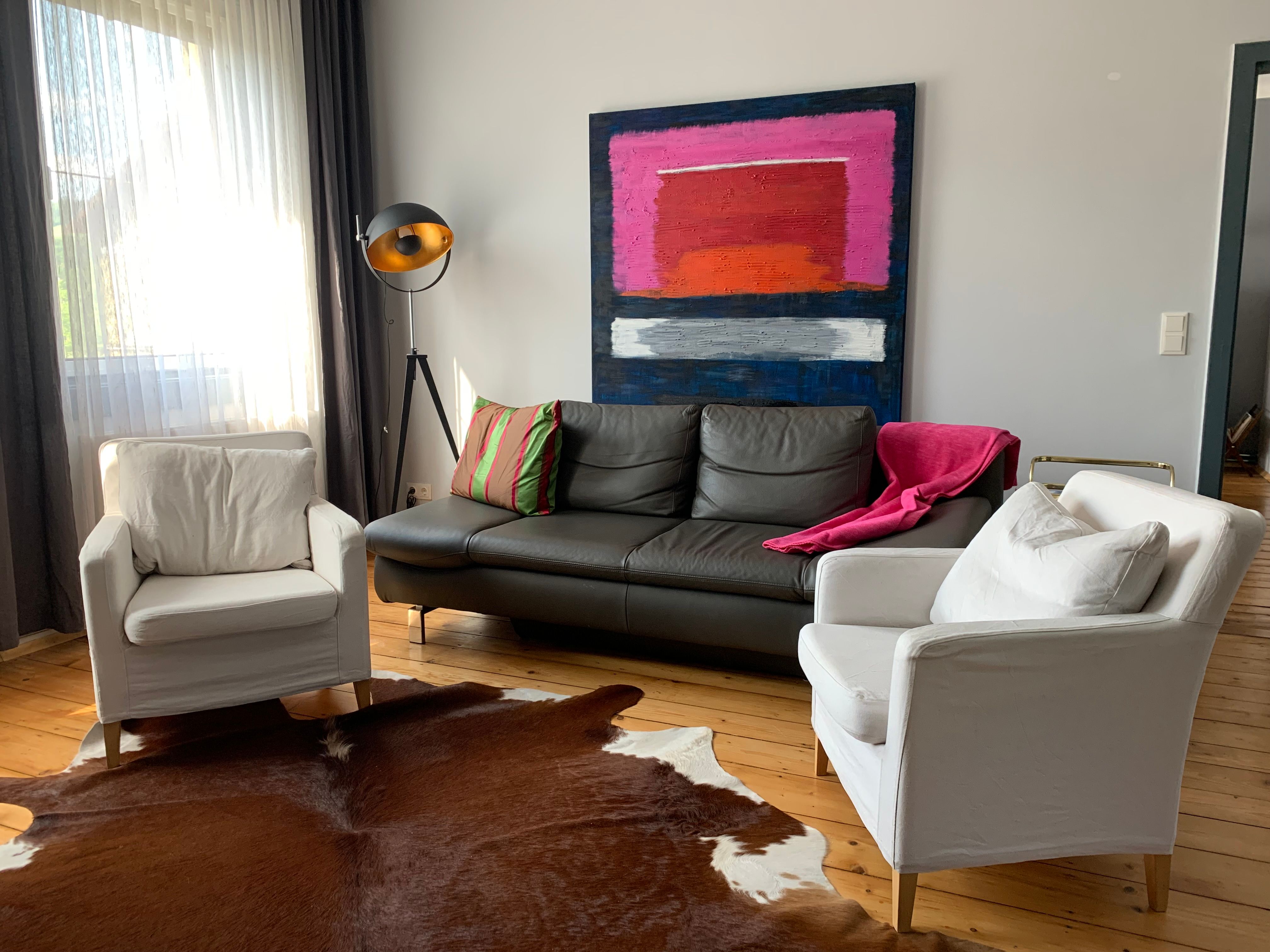 Modernes Wohnzimmer mit Ledersofa, zwei weißen Sesseln, Stehlampe und abstraktem Gemälde an der Wand.