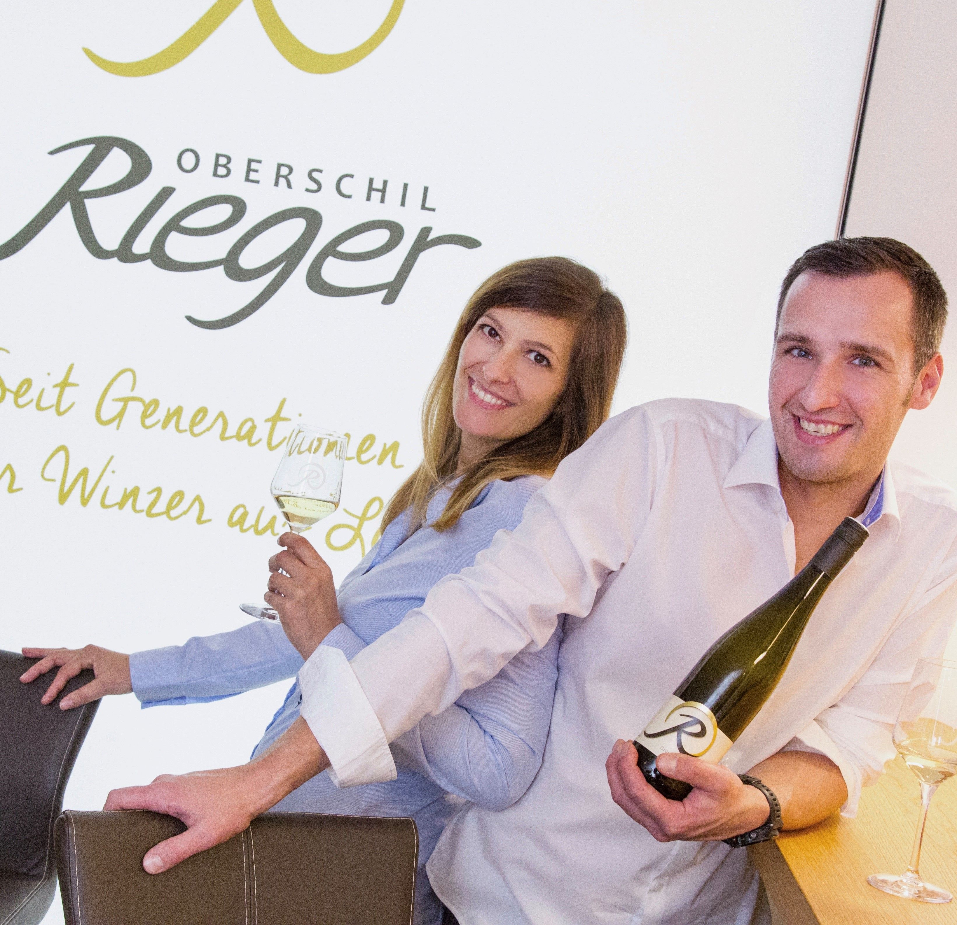 Zwei Personen lächeln und halten Weinflaschen vor einem Schild mit der Aufschrift 'Oberschil Rieger'.