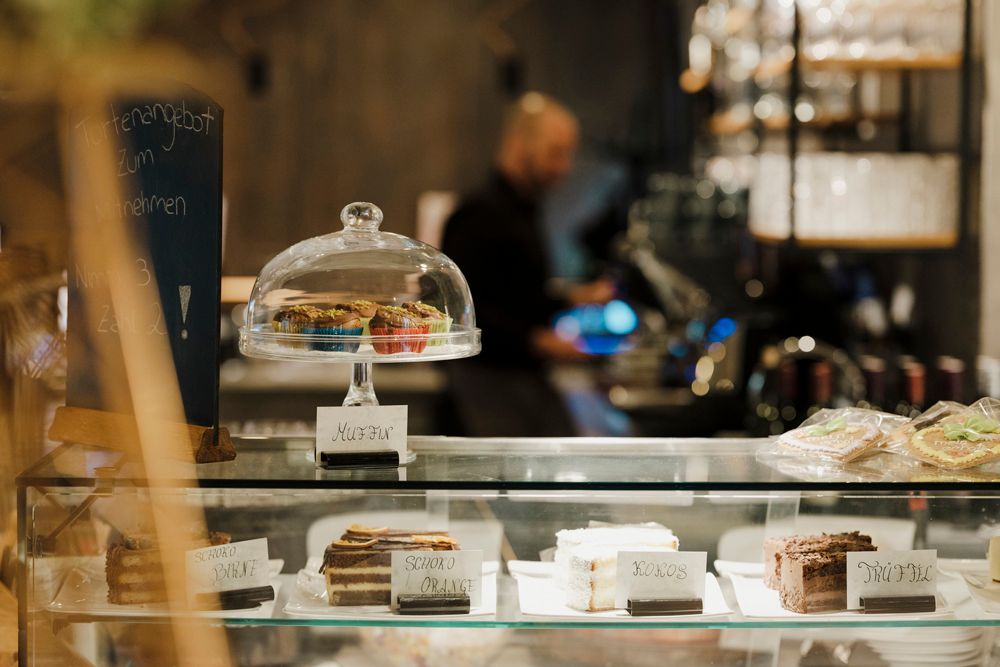 Konditorei mit Kuchen und Muffins in Glasvitrine, Angebotstafel im Hintergrund.