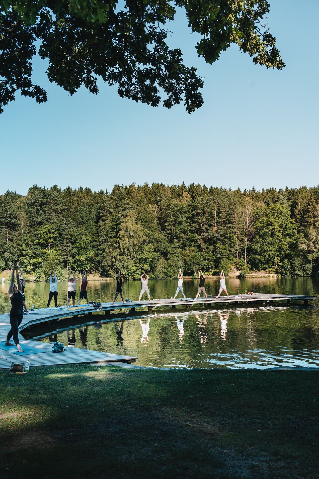 Am Ufer eines ruhigen Sees praktizieren Teilnehmer Yoga und genießen die harmonische Verbindung von Körper und Natur. Umgeben von üppigem Grün und dem sanften Plätschern des Wassers entsteht eine Atmosphäre der Entspannung und inneren Ruhe.