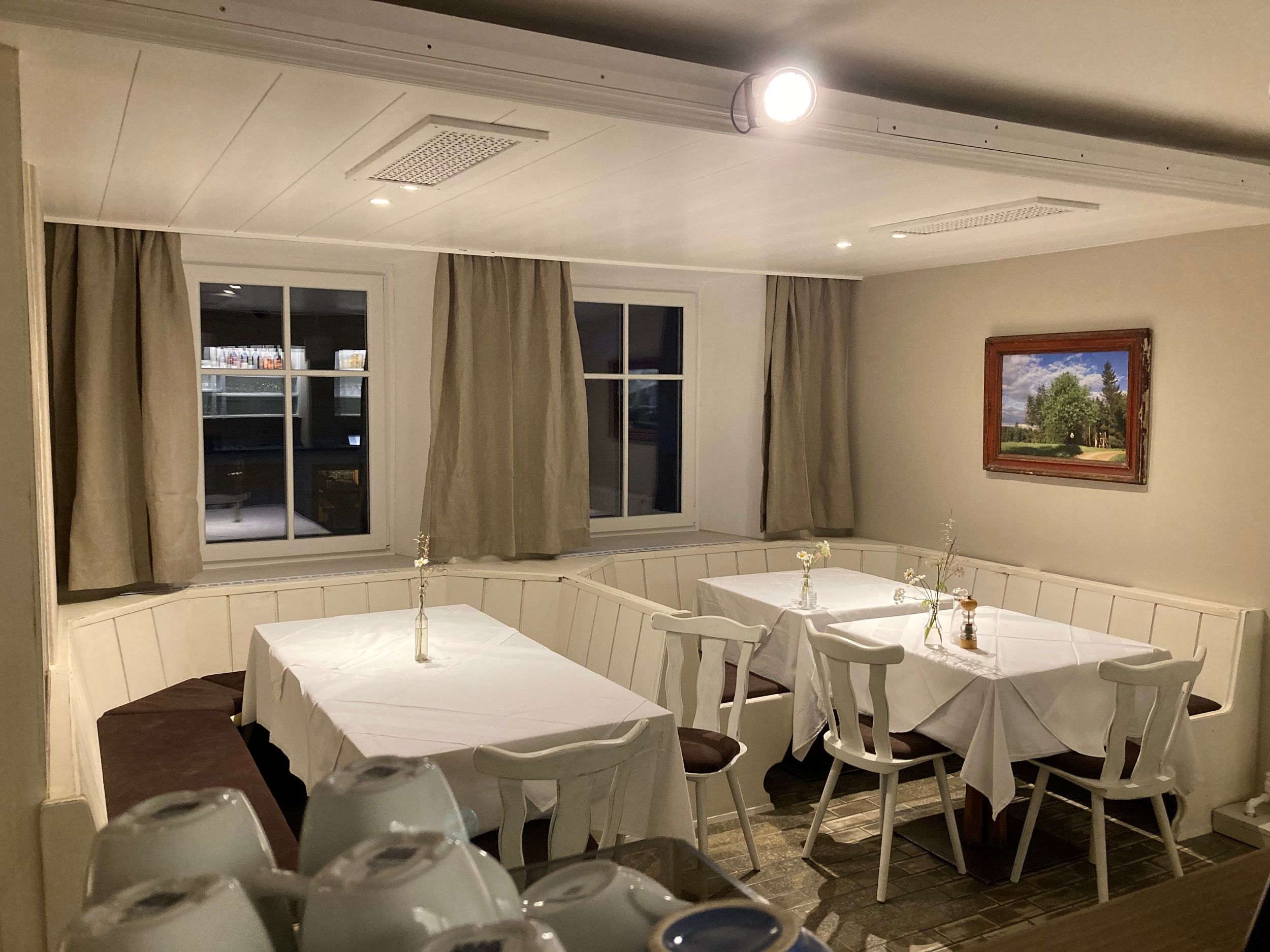 Ein gemütliches Gastzimmer mit zwei gedeckten Tischen, weißen Tischdecken und Stühlen. An der Wand hängt ein Landschaftsbild, und durch die Fenster sind Vorhänge zu sehen.