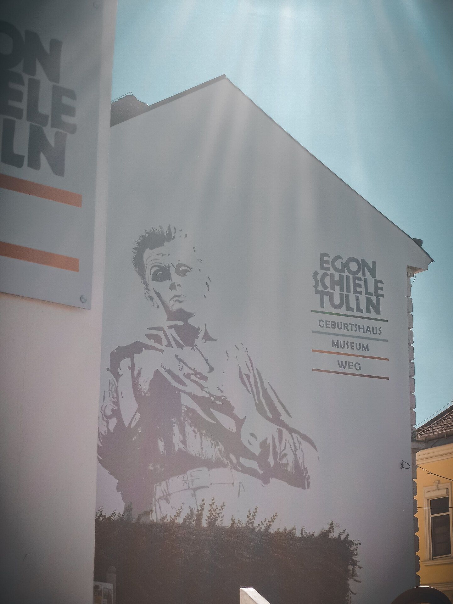 Die strahlende Sonne wirft sanfte Lichtstrahlen auf die beeindruckende Wandmalerei, die den berühmten Künstler in seiner charakteristischen Pose zeigt. Umgeben von der malerischen Kulisse des Herbstes lädt dieser Ort dazu ein, in die kreative Welt von Egon Schiele einzutauchen und die Schönheit der Donau zu genießen.