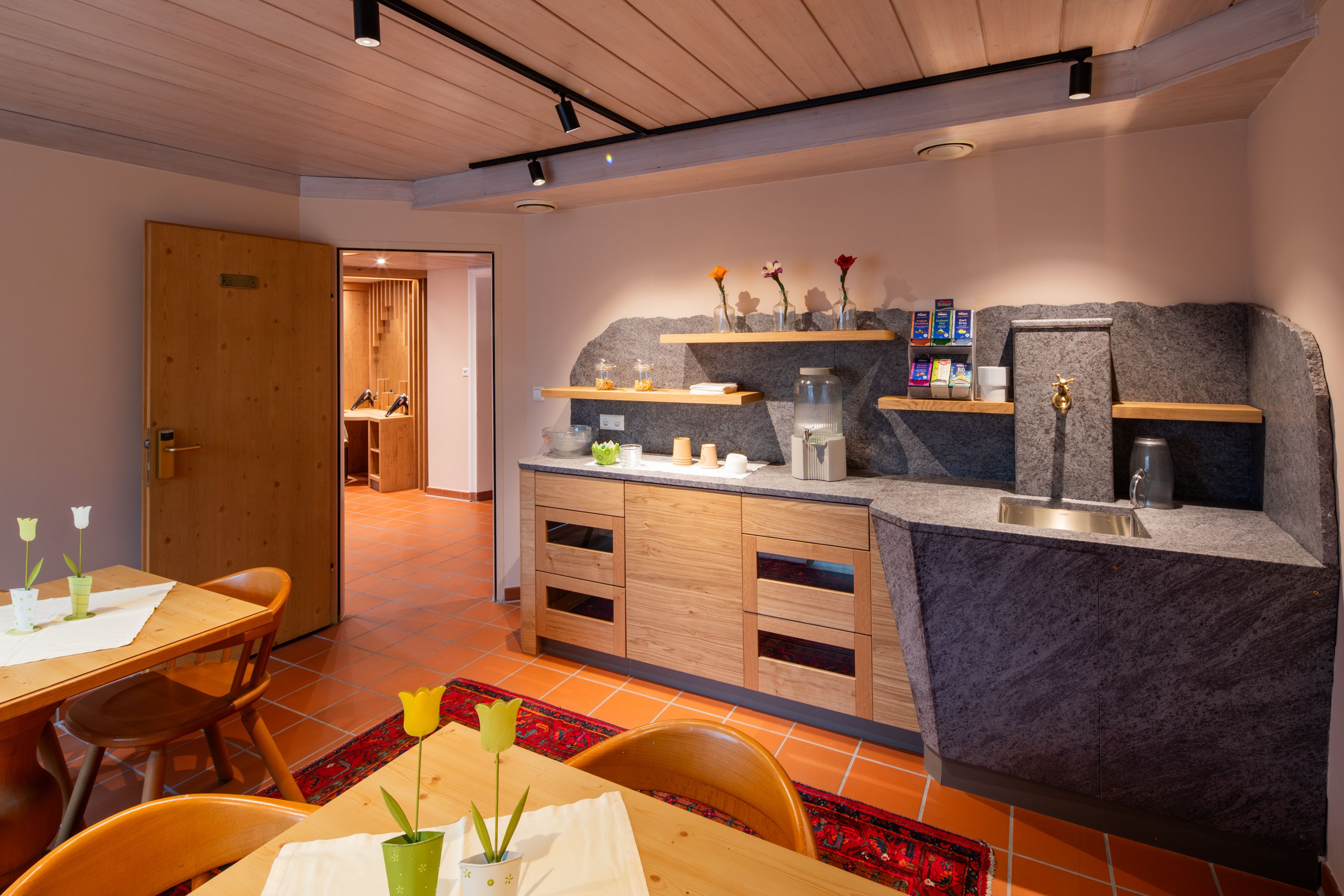 Ein gemütlicher Aufenthaltsraum in einem Hotel-Spa mit Holzmöbeln, einer kleinen Küchenzeile und dekorativen Blumen.