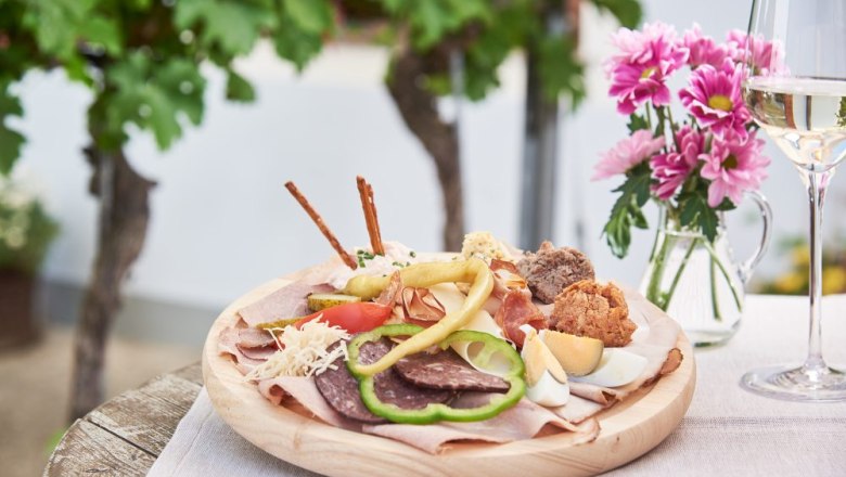 Ein Teller mit verschiedenen Heurigen-Spezialit&auml;ten, daneben ein Glas Wei&szlig;wein und rosa Blumen auf einem Tisch im Freien.