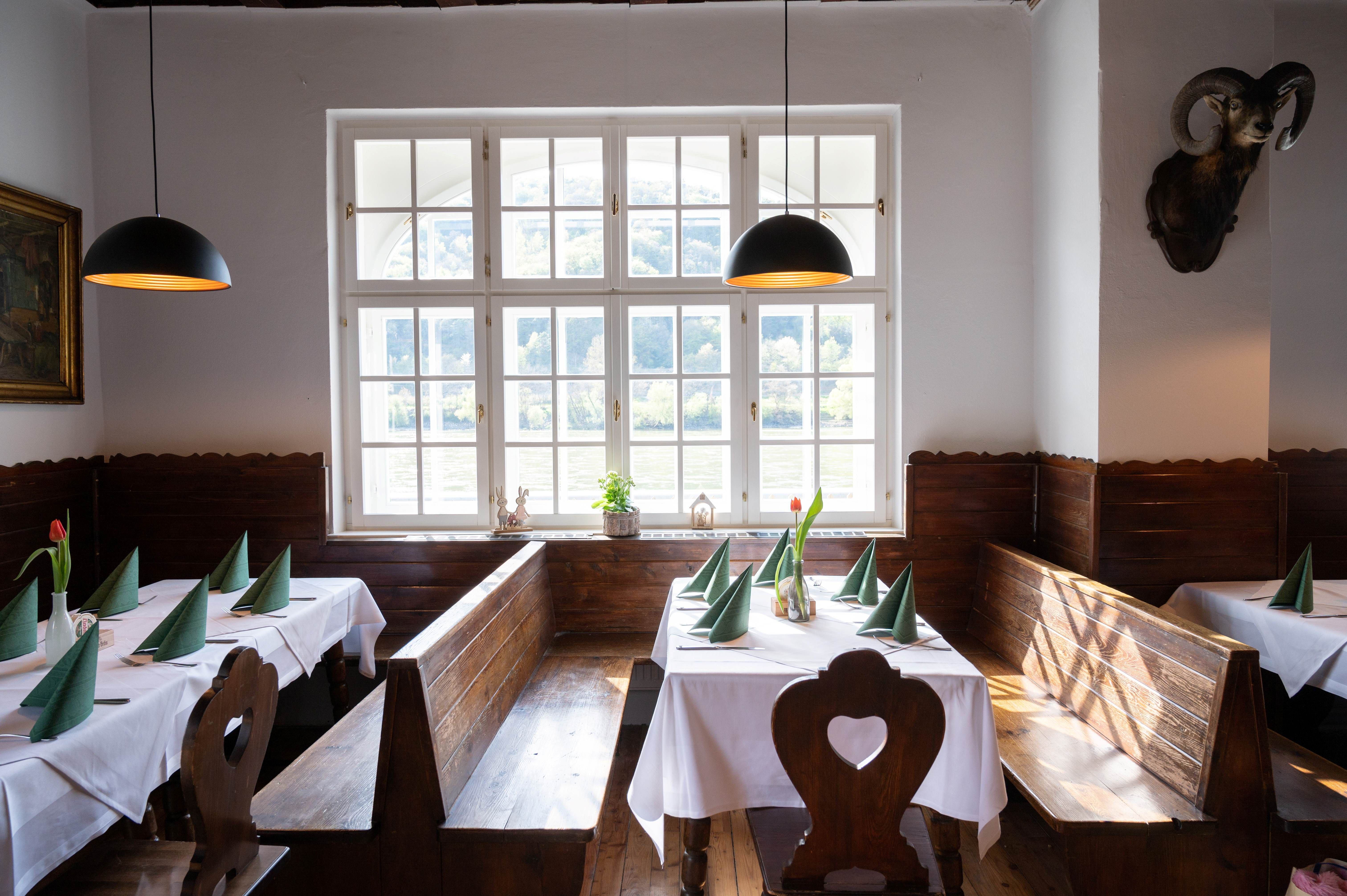 Gemütliche Gaststube mit Holzbänken, weißen Tischdecken und grünen Servietten. Große Fenster mit Blick auf die Natur.