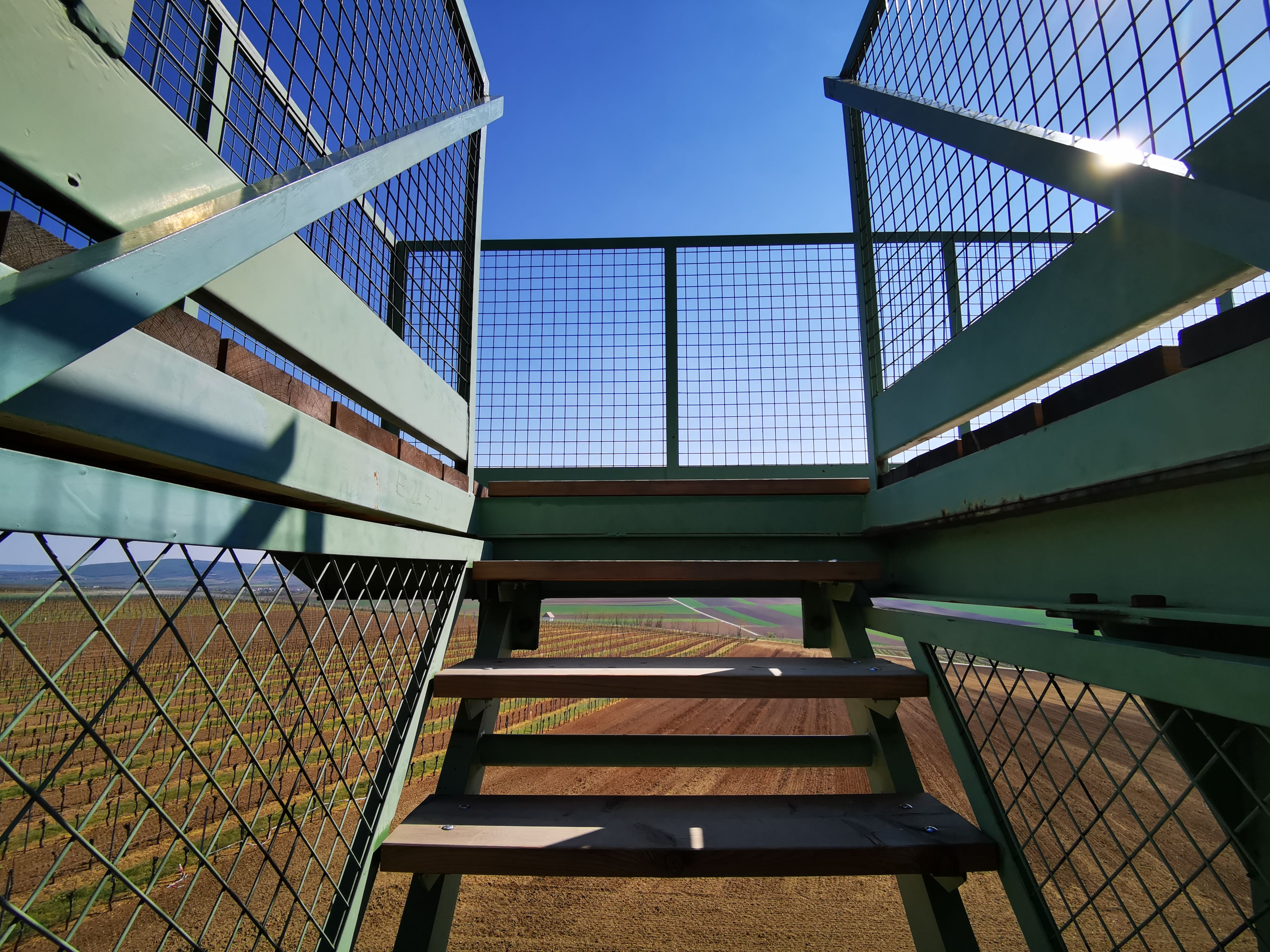 Blick von einer Treppe mit Metallgeländer auf Felder und blauen Himmel.