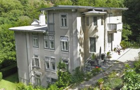 Großes, mehrstöckiges Haus mit grauer Fassade, umgeben von Bäumen und Garten.