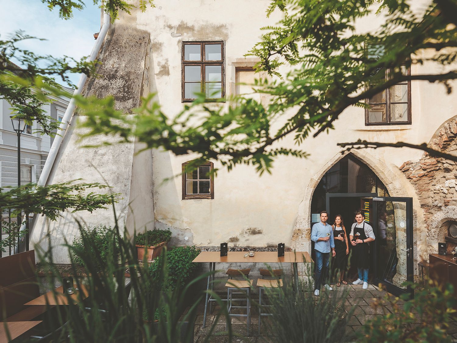 Drei Personen stehen in einem historischen Innenhof vor einem Gebäude mit gotischem Bogenfenster.