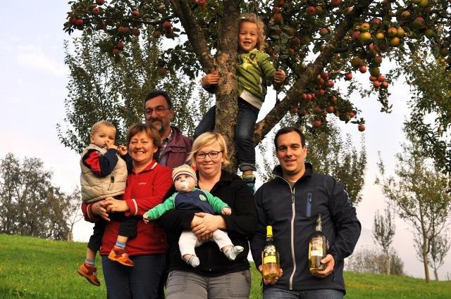 Eine Familie steht vor einem Apfelbaum auf einer Wiese. Zwei Erwachsene halten Kleinkinder, ein Kind sitzt im Baum. Ein Mann hält zwei Flaschen.