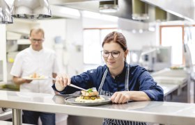 K&ouml;chin in einer Restaurantk&uuml;che richtet ein Gericht an, w&auml;hrend ein Kollege im Hintergrund ein weiteres Gericht h&auml;lt.