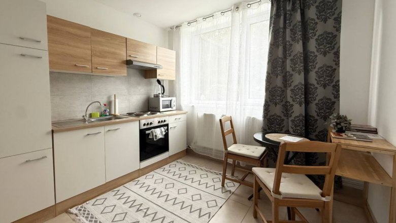 Kleine K&uuml;che mit Holzm&ouml;beln, Esstisch und St&uuml;hlen, helle Vorh&auml;nge und Teppich.