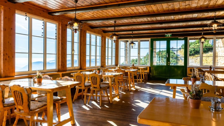 Gem&uuml;tliche Gaststube mit Holzm&ouml;beln und gro&szlig;en Fenstern, die einen Blick auf die Berge bieten.