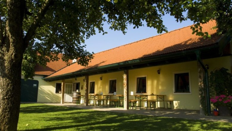 Ein Heurigengarten mit gelbem Geb&auml;ude, roten Dachziegeln und gr&uuml;ner Wiese.