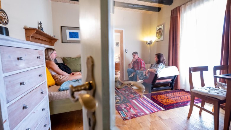 Drei Personen sitzen in einem gemütlichen Wohnzimmer mit Holzboden und buntem Teppich.