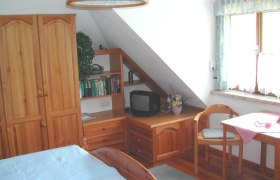 Gemütliches Gästezimmer mit Holzmöbeln, einem kleinen Fernseher, einem Tisch mit Stuhl und einem Fenster mit Vorhängen.