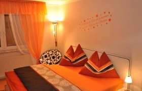 Gemütliches Schlafzimmer mit orangefarbener Bettwäsche und Vorhängen, dekorativen Kissen und Wandtattoo.
