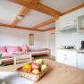 Gem&uuml;tliche Ferienwohnung mit Holzm&ouml;beln, einer kleinen K&uuml;che und einem Bett mit rot-wei&szlig; karierten Kissen. Ein Obstkorb steht auf dem Tisch.