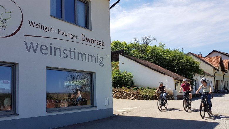 Drei Personen fahren mit Fahrr&auml;dern an einem Geb&auml;ude mit der Aufschrift 'Weingut - Heuriger - Dworzak weinstimmig' vorbei.