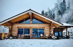 Blockh&uuml;tte im Schnee mit gro&szlig;en Fenstern und Terrasse, umgeben von verschneiten B&auml;umen.