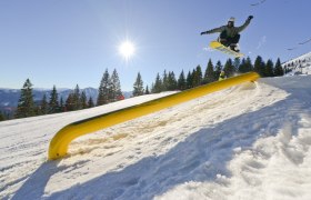 Snowboarder springt &uuml;ber ein gelbes Hindernis im Snowpark Gemeindealpe bei Sonnenschein.