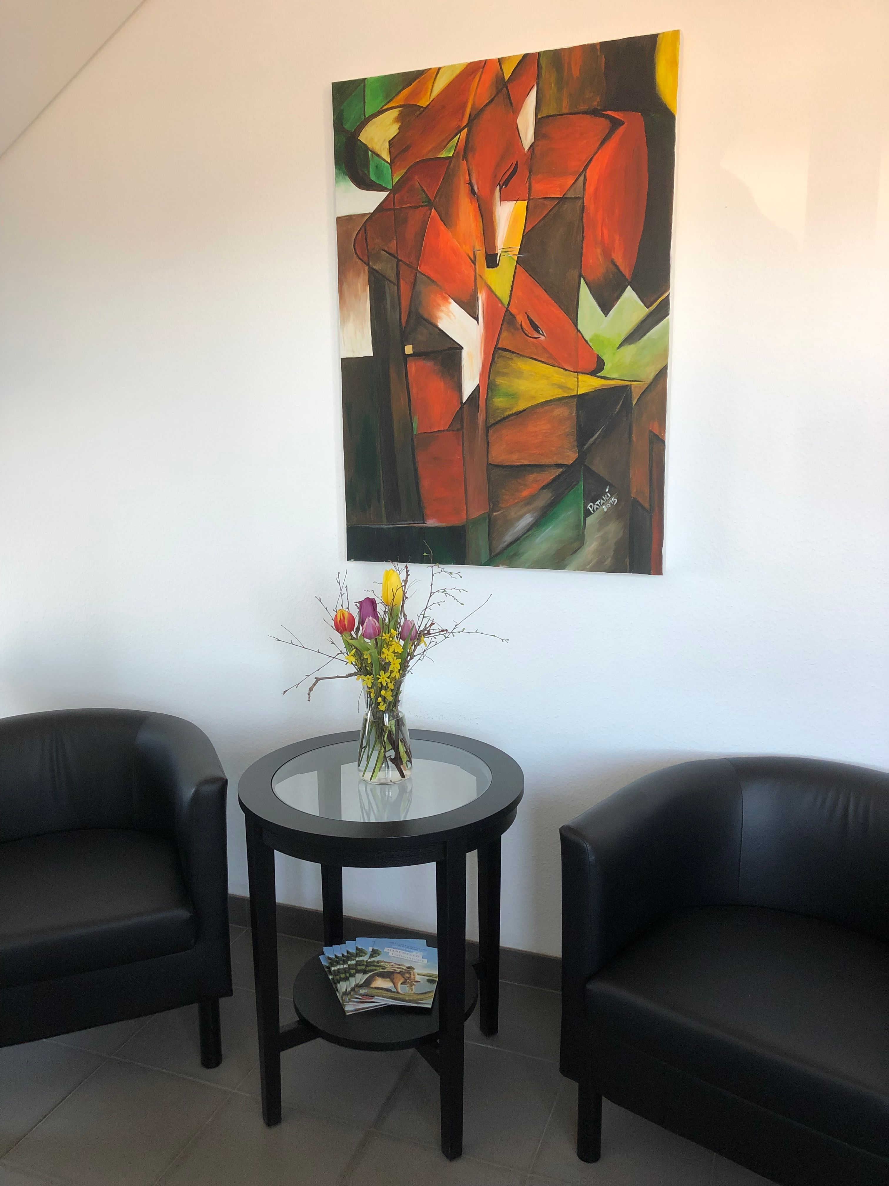 Zwei schwarze Sessel und ein runder Tisch mit Blumen in einem Raum. An der Wand hängt ein abstraktes Gemälde mit Fuchs-Motiv.