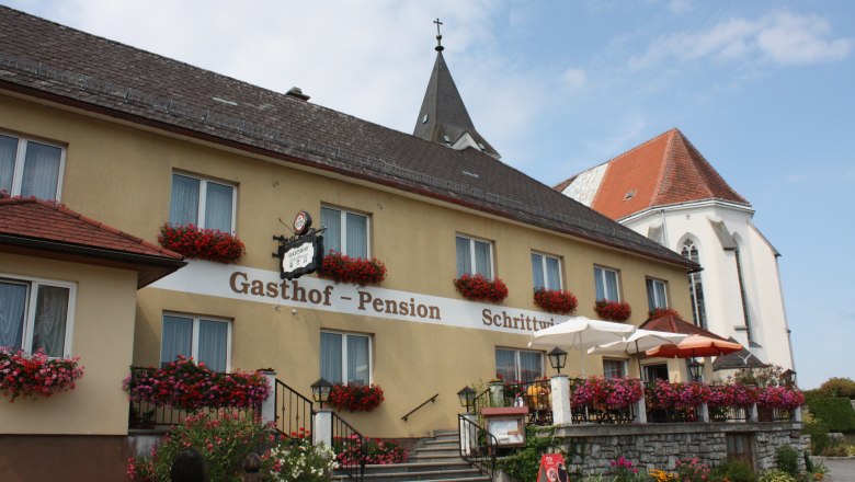 Gasthof Schrittwieser, © zVg Gemeinde Texing Ein traditioneller Gasthof mit Blumen geschmückter Fassade und Terrasse, daneben eine Kirche mit spitzem Turm.