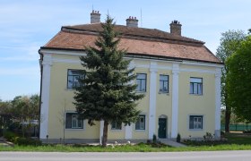 Gelbes zweistöckiges Gebäude mit rotem Ziegeldach und grüner Tür, umgeben von Bäumen und einem kleinen Garten.