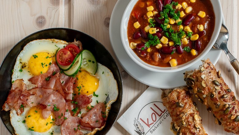 Verschiedene warme Snacks auf einem Tisch, darunter Spiegeleier mit Speck, Chili con Carne, belegte Brote und Br&ouml;tchen.