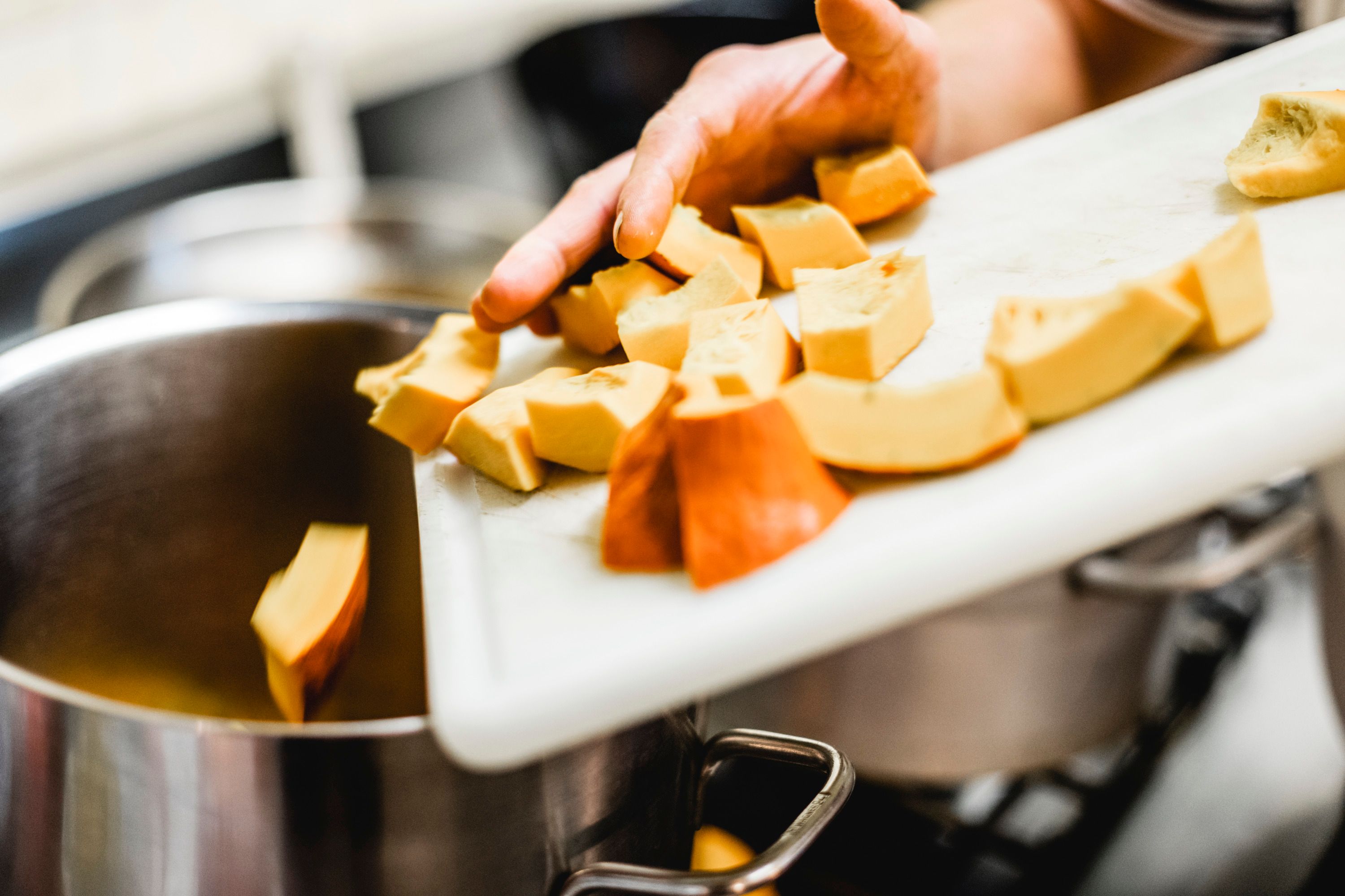 Eine Hand schiebt geschnittene Kürbisstücke von einem Schneidebrett in einen Topf.
