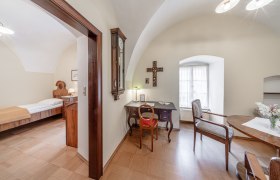 Ein traditionell eingerichtetes Zimmer mit einem Schreibtisch, Stuhl, Tisch und Bett. Ein Kreuz h&auml;ngt an der Wand, und ein Fenster l&auml;sst Tageslicht herein.