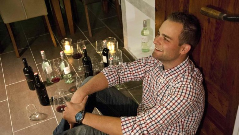 Ein Mann sitzt l&auml;chelnd auf dem Boden, umgeben von Weinflaschen und Gl&auml;sern.