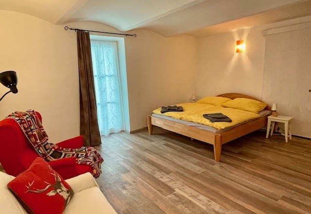 Gem&uuml;tliches Schlafzimmer mit Holzboden, Doppelbett, rotem Sessel und Fenster mit Vorhang.