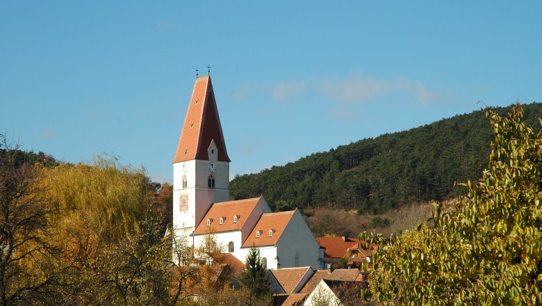 Kirche in Nu&szlig;dorf mit rotem Dach und Uhrturm, umgeben von B&auml;umen und H&uuml;geln.