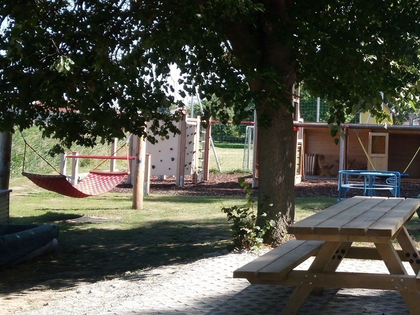 Ein Spielplatz mit einer Hängematte, Kletterwand und Holztisch unter einem Baum.