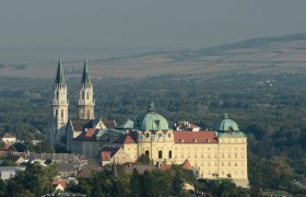 Luftaufnahme des Stifts Klosterneuburg mit zwei Türmen und einer Kuppel, umgeben von Wald und Feldern.