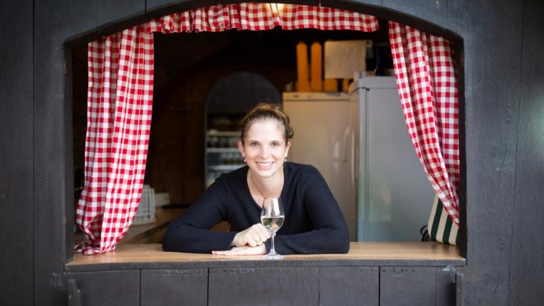 Frau lehnt sich aus einem Fenster mit rot-wei&szlig; karierten Vorh&auml;ngen, h&auml;lt ein Glas Wein.