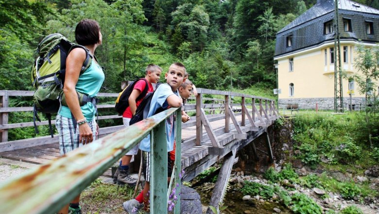 Eine Gruppe von Menschen steht auf einer Holzbr&uuml;cke in einer gr&uuml;nen, bewaldeten Umgebung. Im Hintergrund ist ein gelbes Geb&auml;ude zu sehen.