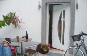 Ein offener Hauseingang mit Tisch, St&uuml;hlen, Weinflasche und Fahrrad.