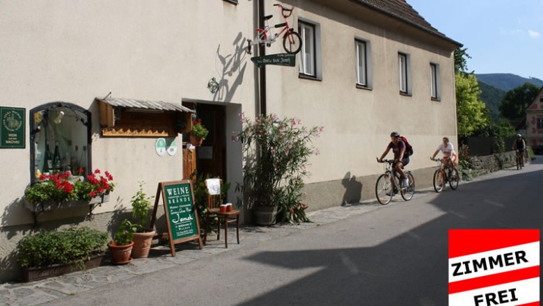 Au&szlig;enansicht eines Hauses mit Fahrraddekoration und Weinausschank. Zwei Radfahrer fahren vorbei. Schild mit 'Zimmer frei'.