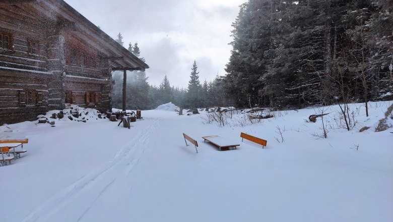 Verschneite Alm mit Holzh&uuml;tte und B&auml;nken im Winterwald.