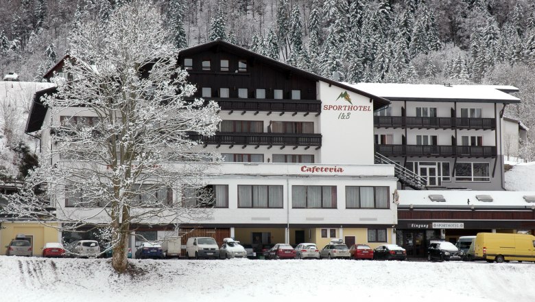 Sporthotel am Ötscher, © Iby & Bogi Matayas Touristik GmbH Ein verschneites Hotelgebäude mit der Aufschrift 'Sporthotel' und 'Cafeteria', umgeben von schneebedeckten Bäumen und geparkten Autos.