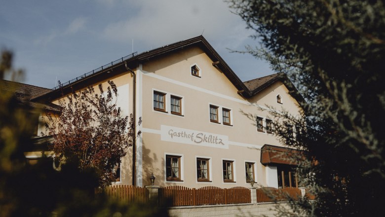 Ein traditionelles Gasthaus mit der Aufschrift 'Gasthof Skilitz', umgeben von Bäumen und einem Holzzaun.
