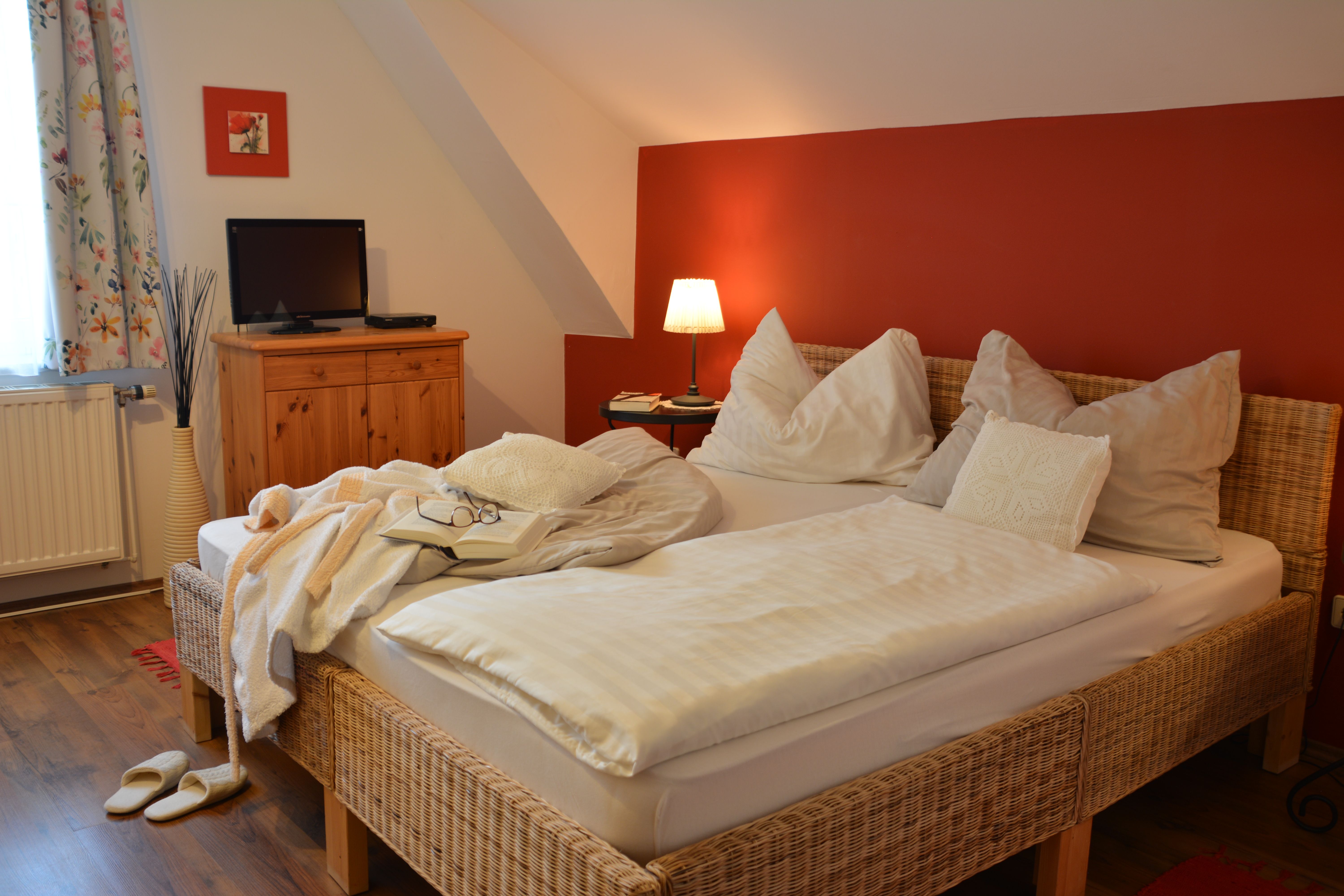 Gemütliches Doppelzimmer mit roter Wand, Bett, Nachttischlampe, Fernseher und Holzmöbeln.
