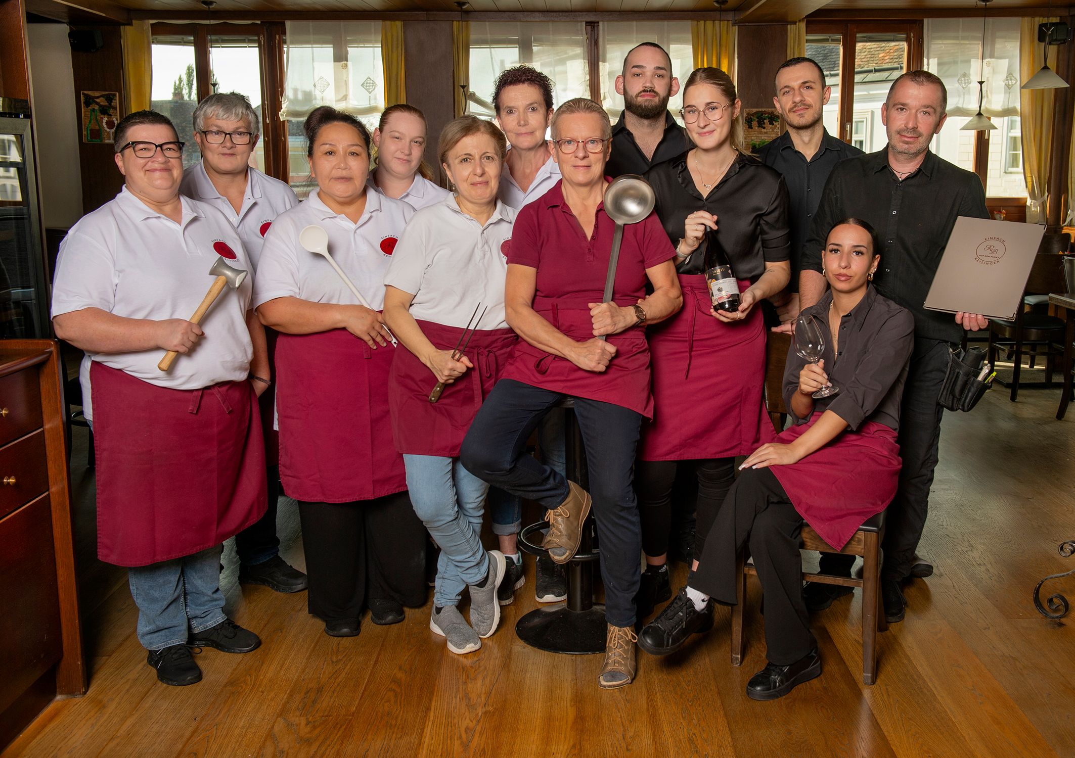 Ein Team von Restaurantmitarbeiterinnen und Mitarbeitern posiert in Arbeitskleidung mit Küchenutensilien in einem Speiseraum.