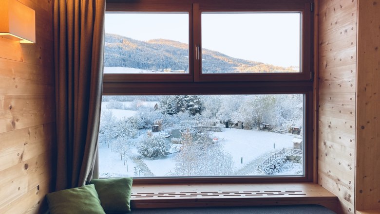 Blick aus einem Holzfenster auf eine verschneite Landschaft mit Bergen im Hintergrund.