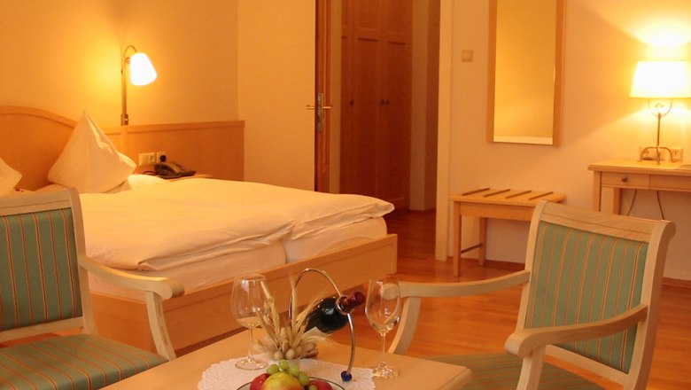 Ein gemütliches Hotelzimmer mit Doppelbett, zwei Stühlen, einem Tisch mit Obst und Wein, und warmem Licht.