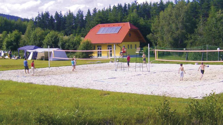 Menschen spielen Volleyball auf einem Sandplatz vor einem gelben Geb&auml;ude mit rotem Dach, umgeben von B&auml;umen.