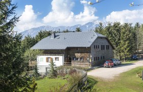 Ein rustikales Schutzhaus in einer bergigen Landschaft mit Bäumen und Autos im Vordergrund.